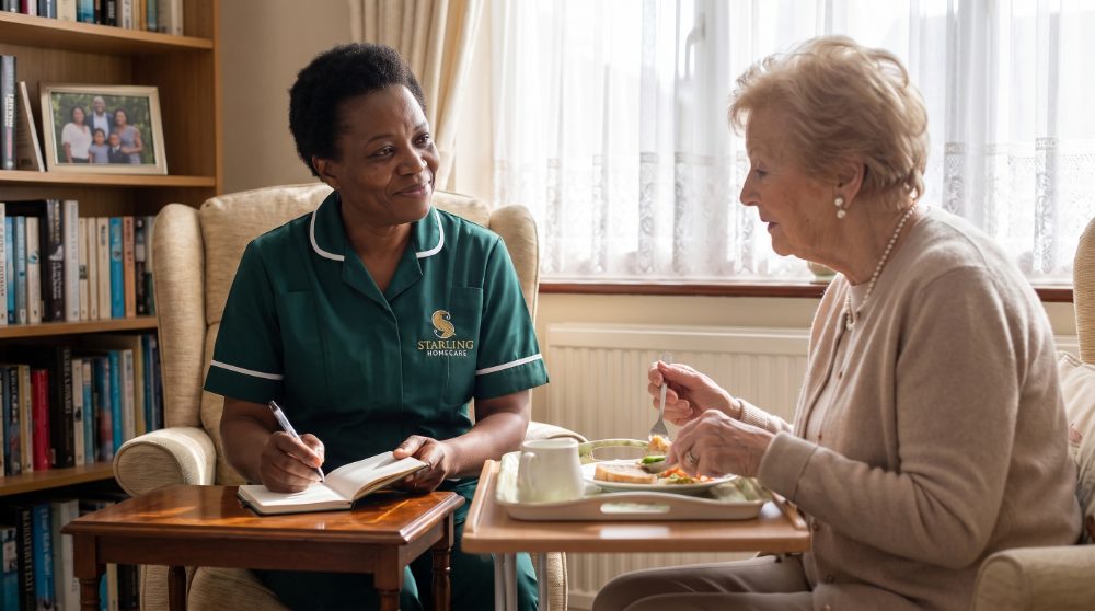 A Starling Homecare carer writing a visit note beside an older woman eating lunch during a daytime dementia care visit in Potters Bar