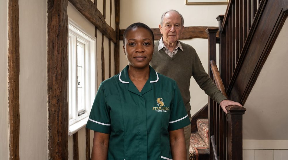 A Starling Homecare carer at the foot of a narrow period staircase in Old Hatfield, with the client standing independently holding the banister
