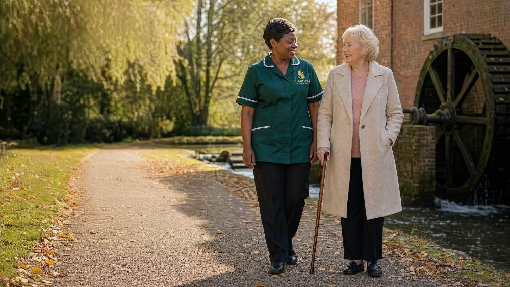 Female carer in bottle green uniform walking with an older woman by a watermill near Hatfield, companionship care