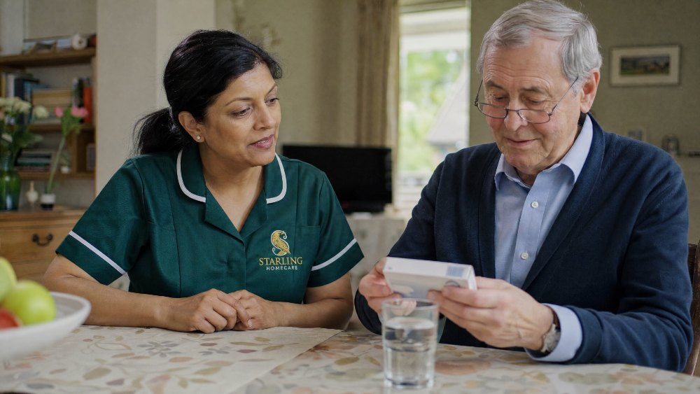 Carer sitting with an older man at his kitchen table as he looks at his medication, a calm conversation about his tablets